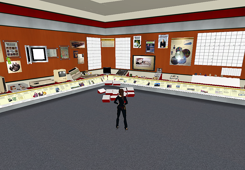Wide interior view of the Avnet Technology Museum in Second Life with an avatar standing center floor surrounded by wall-mounted displays, video screens, exhibit cases, and build component boxes, recreating the layout of Avnet's Phoenix corporate lobby. The image shows the project under construction.